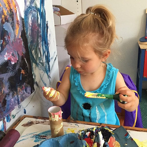 6 reasons to teach a toddler to paint - girl with paint brush in one hand, used to paint the other hand. Background wall has been painted in various colours by the toddler.